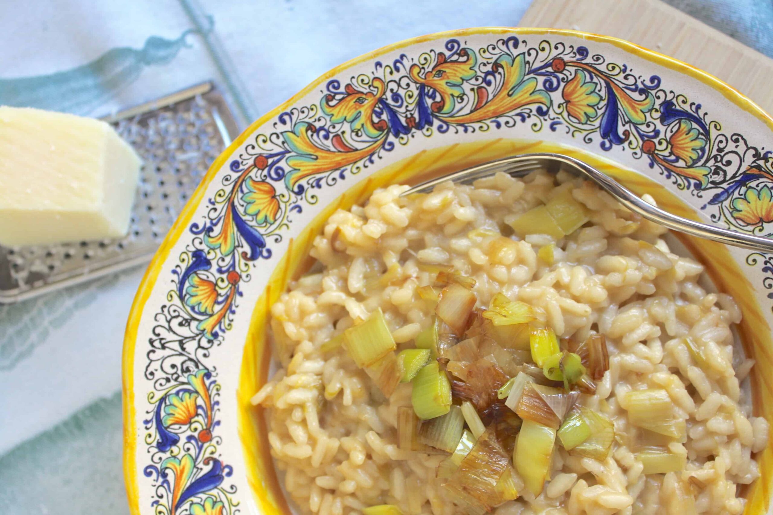 A bowl of risotto topped with sautéed leeks, next to a metal grater and a block of cheese on a patterned plate.