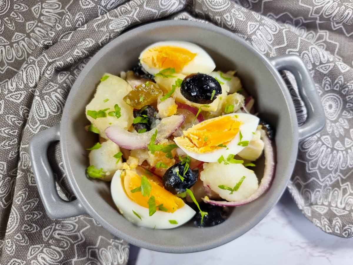 A bowl of potato salad with sliced hard-boiled eggs, black olives, red onion, parsley, and green olives, placed on a patterned cloth.