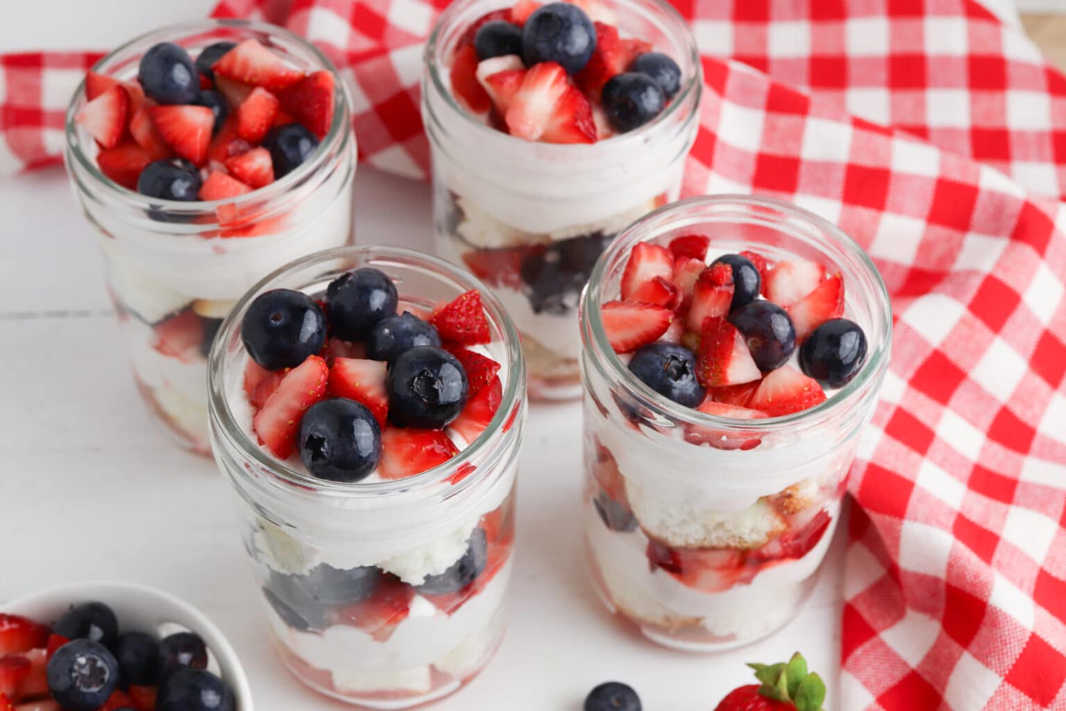 Four mason jars filled with layered dessert of whipped cream, sponge cake, strawberries, and blueberries, set on a white surface with a red and white checkered cloth.