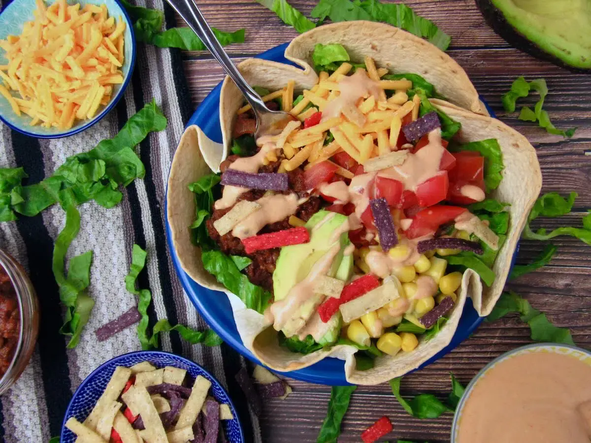 A tortilla bowl filled with lettuce, tomatoes, avocado, corn, tortilla strips, shredded cheese, and creamy dressing, surrounded by more toppings on a wooden surface.