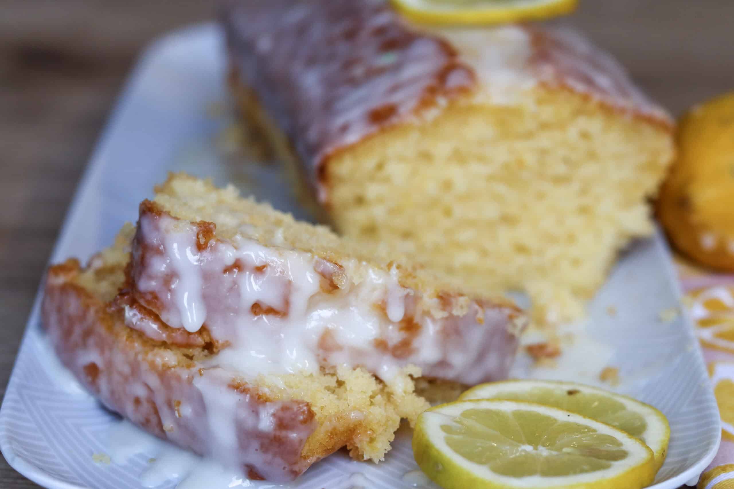 A loaf of lemon cake with icing is sliced on a white plate, with lemon slices placed nearby.