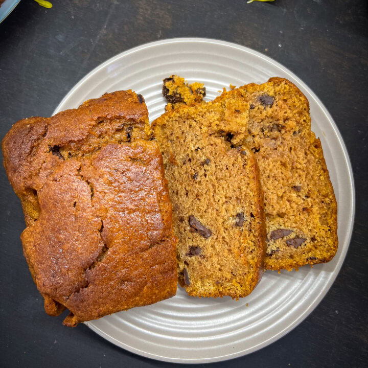 A loaf of banana bread with two slices cut, showing a moist texture and dark chocolate chunks, served on a beige plate.