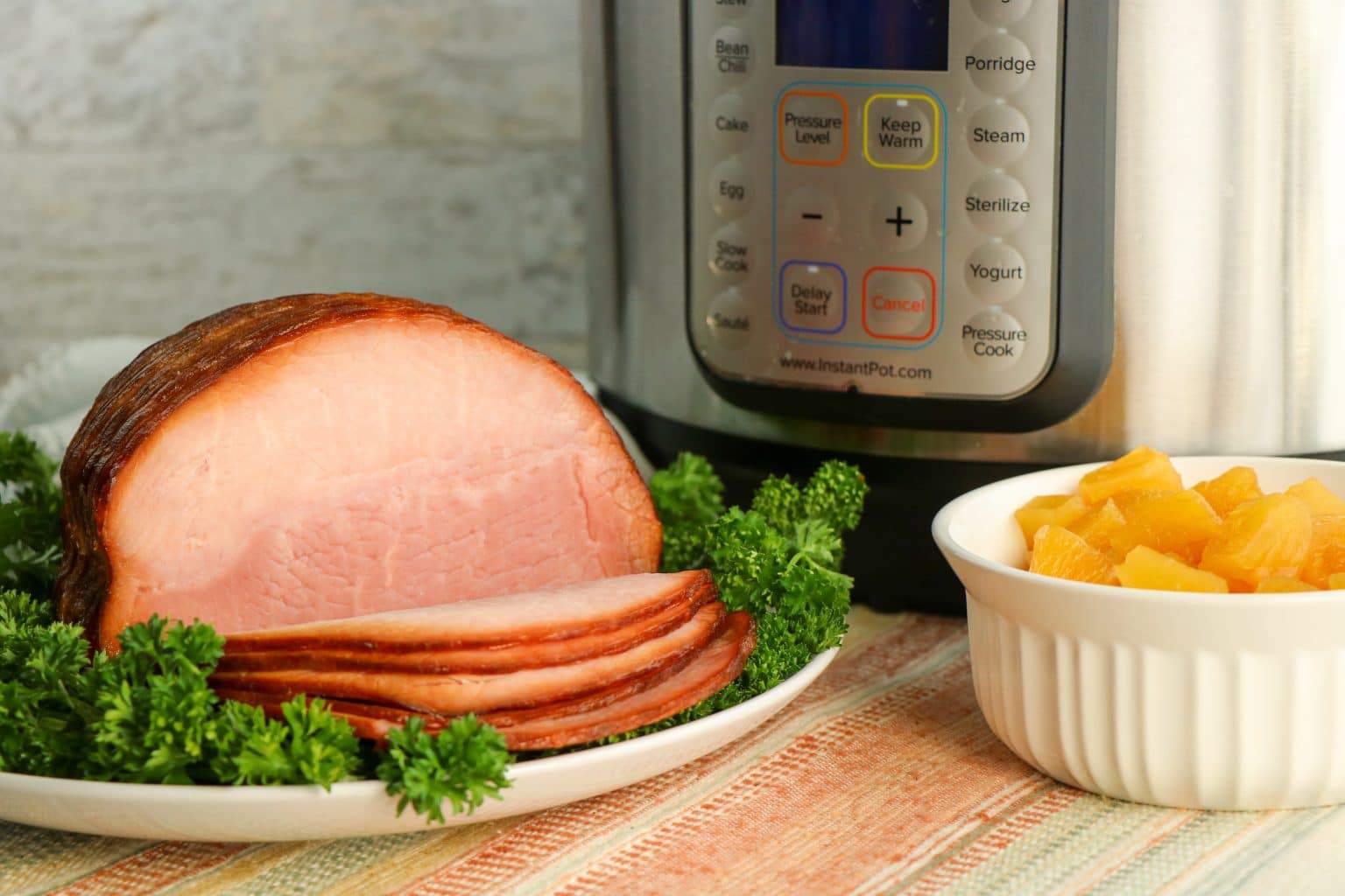 A sliced ham on a plate with parsley, a bowl of pineapple chunks, and an Instant Pot in the background.