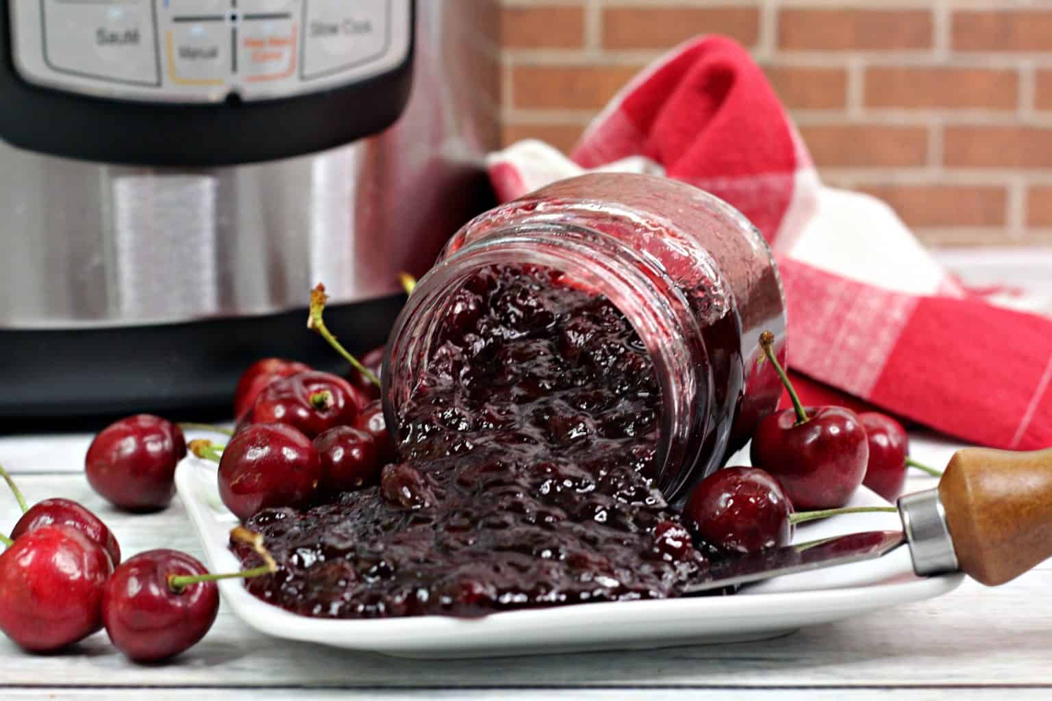A glass jar of cherry jam tipped over on a white plate, surrounded by fresh cherries, with a knife and an Instant Pot in the background.