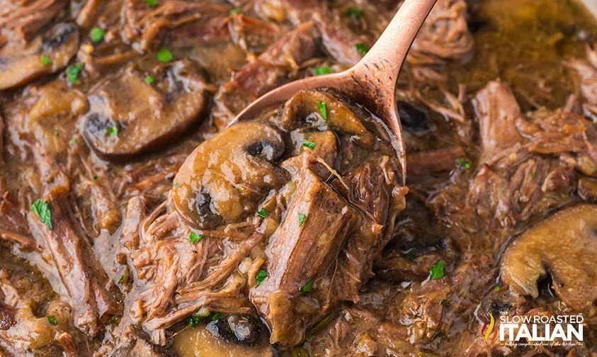 Close-up of shredded beef with sliced mushrooms in a brown gravy, served with a spoon.