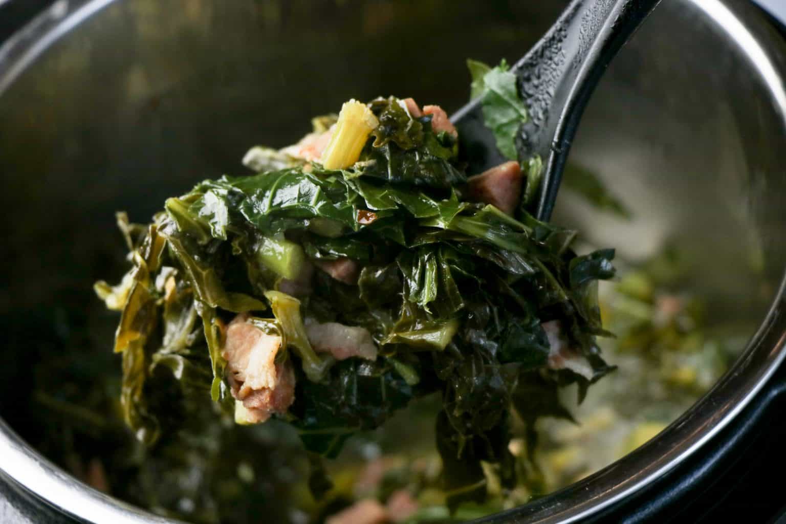 A close-up of cooked collard greens mixed with pieces of meat, being lifted from a pot with a black serving spoon.