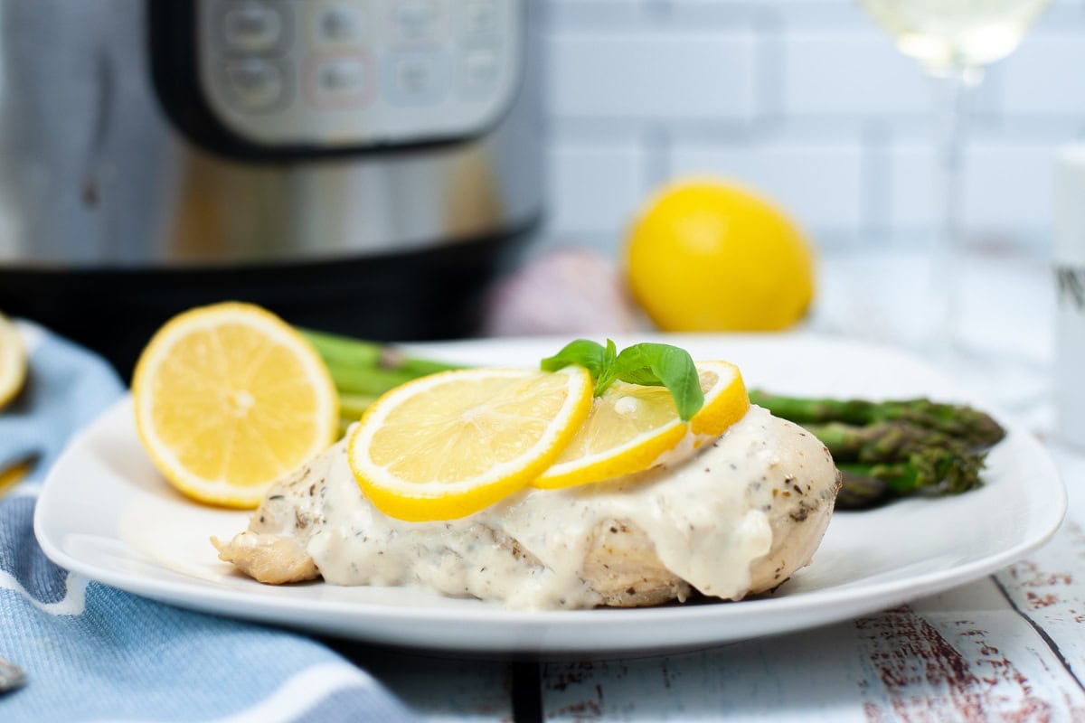 A plate of chicken breast topped with creamy white sauce and lemon slices, served with asparagus; an Instant Pot is visible in the background.