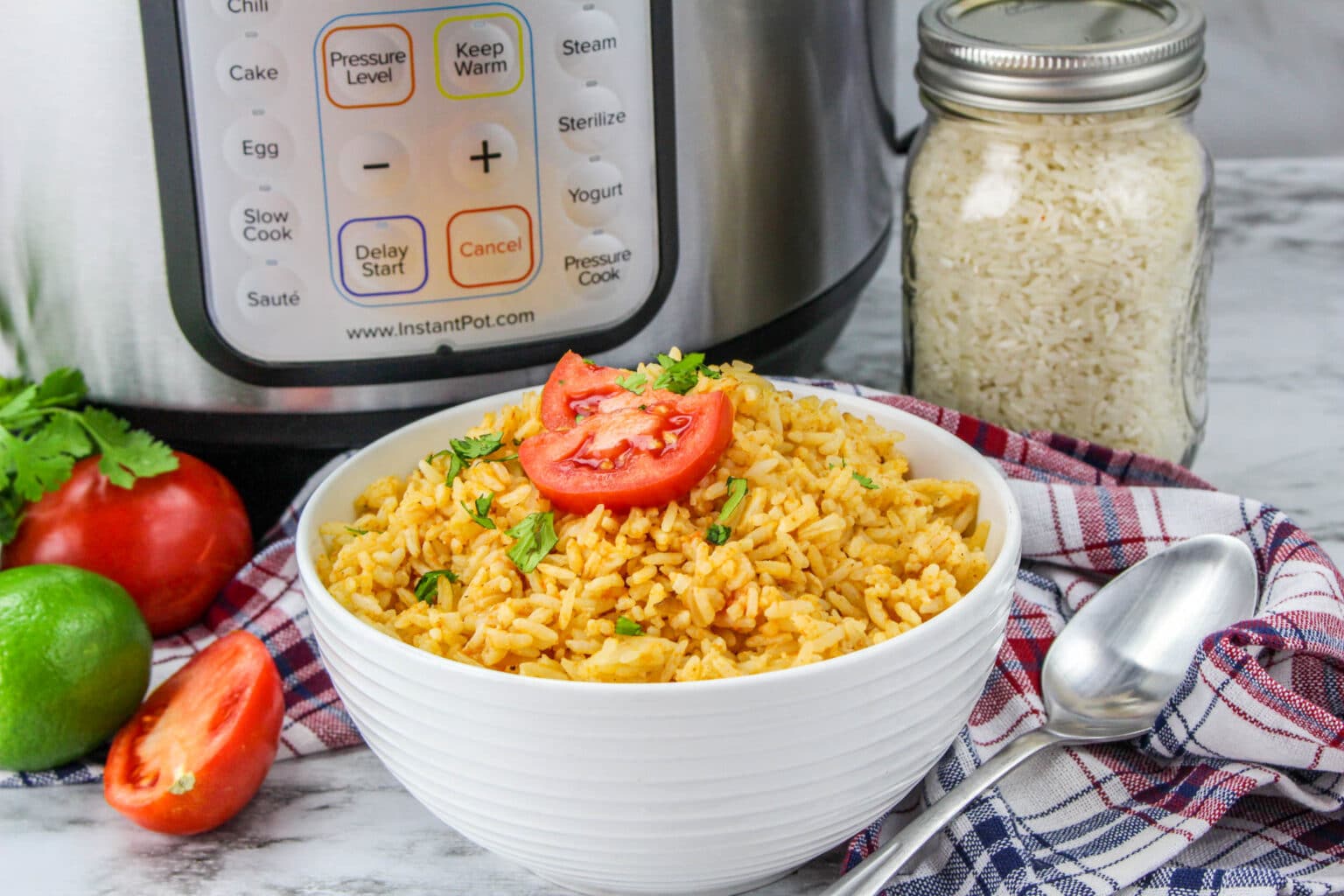 A bowl of seasoned rice topped with tomato and herbs sits in front of an Instant Pot, with a jar of rice, fresh tomato, lime, and a spoon nearby.