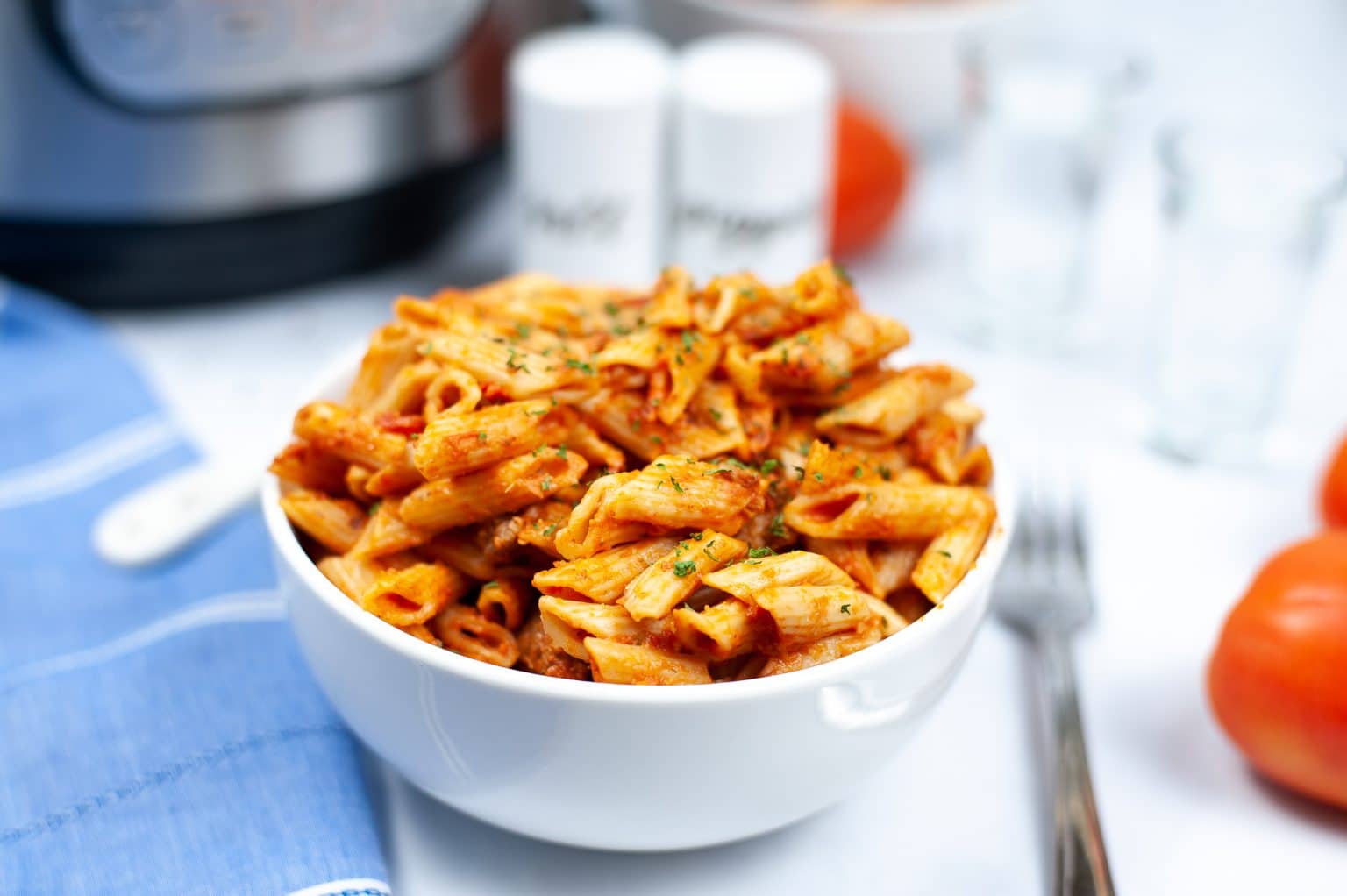 A white bowl filled with penne pasta in tomato sauce, garnished with herbs, sits on a white surface with a fork, tomatoes, and a blue cloth nearby.
