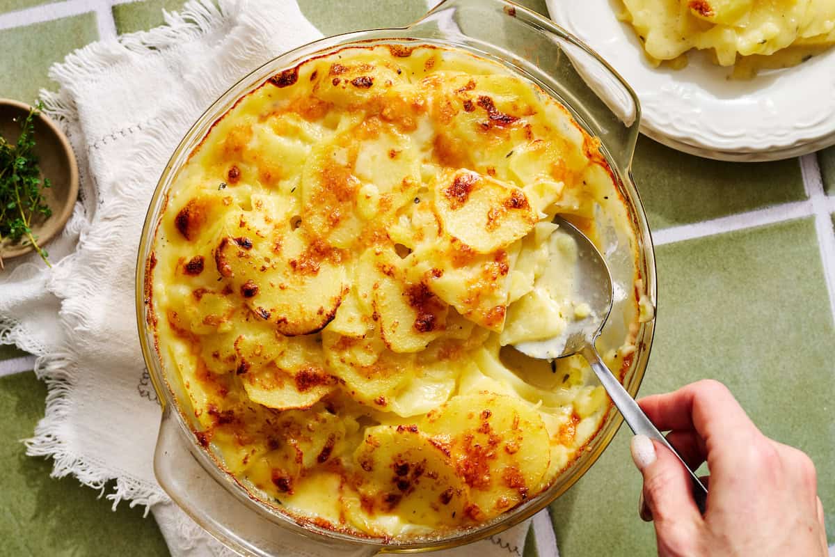 A hand scoops a serving of cheesy, baked scalloped potatoes from a round glass dish on a green tiled surface.