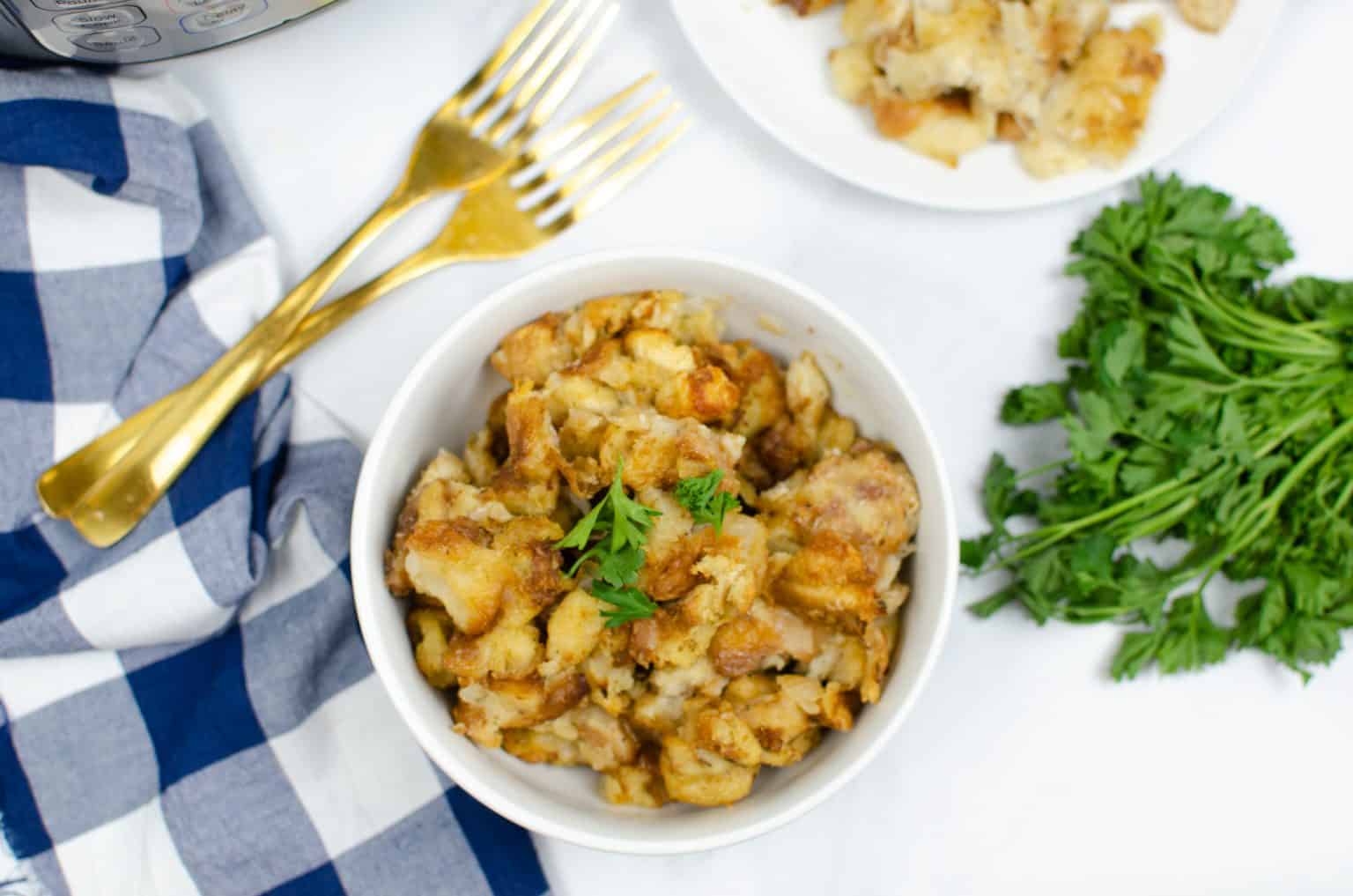 A bowl of bread stuffing garnished with parsley, placed on a white surface with gold forks, a blue and white napkin, and a bunch of fresh parsley nearby.