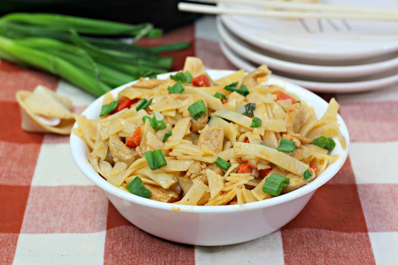 A white bowl filled with stir-fried noodles, tofu, vegetables, and topped with chopped green onions sits on a red and white checkered tablecloth.
