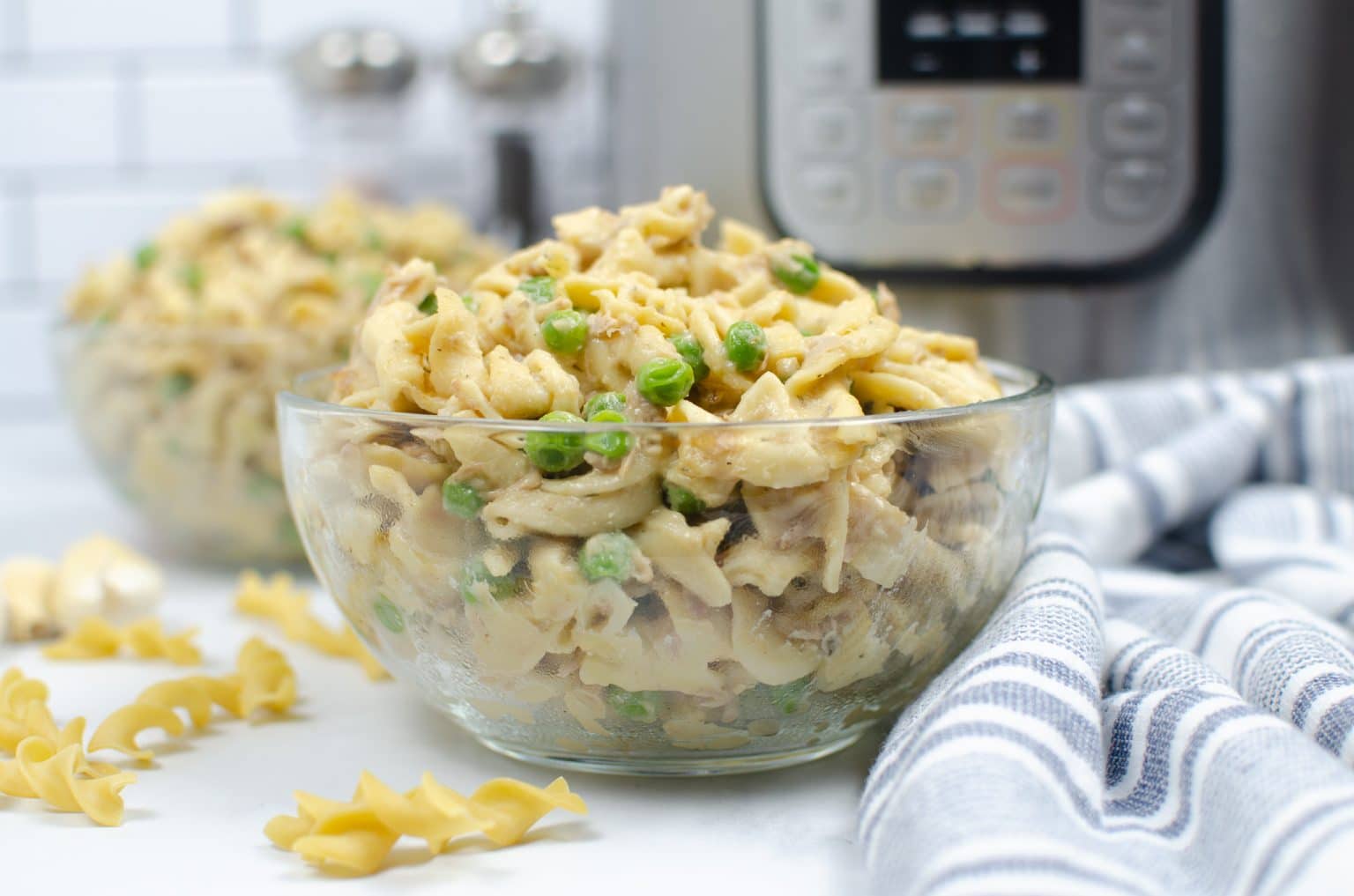 A glass bowl filled with creamy pasta, green peas, and shredded chicken sits on a white surface near a striped towel and a pressure cooker.