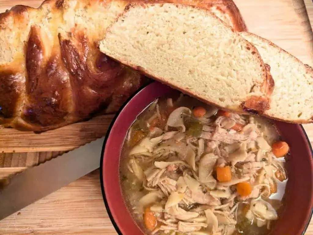 A bowl of chicken noodle soup with carrots and noodles next to two thick slices of braided bread on a wooden cutting board.
