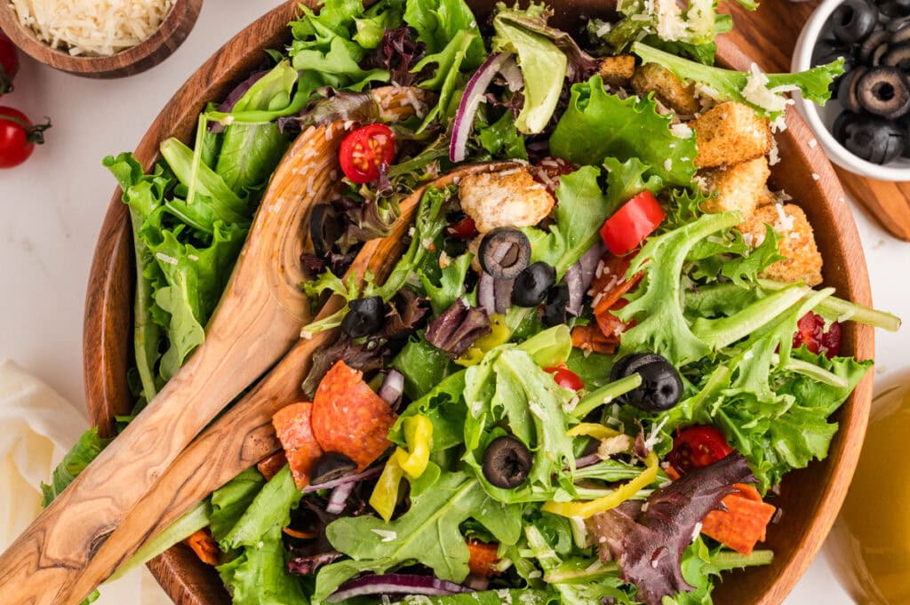 A wooden bowl filled with fresh salad greens, cherry tomatoes, black olives, pepperoni slices, croutons, and shredded cheese, with wooden salad tongs.