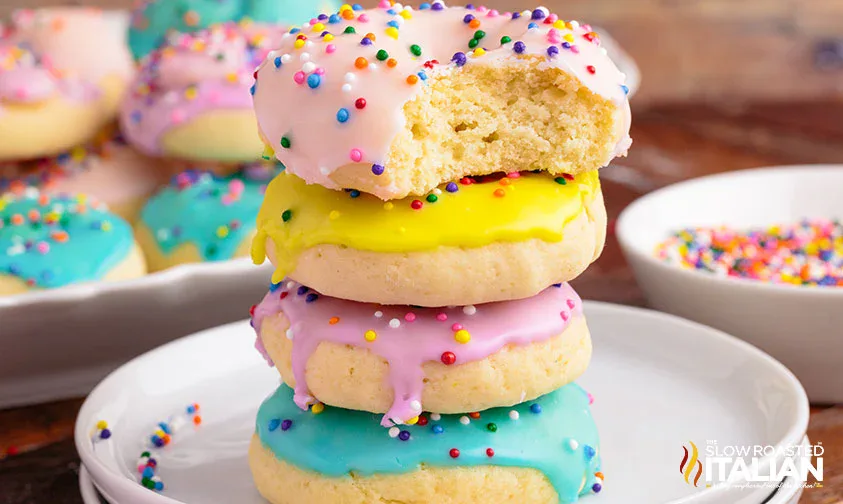 A stack of three frosted cookies with sprinkles, topped with a fourth cookie that has a bite taken out. More frosted cookies and a bowl of sprinkles are in the background.