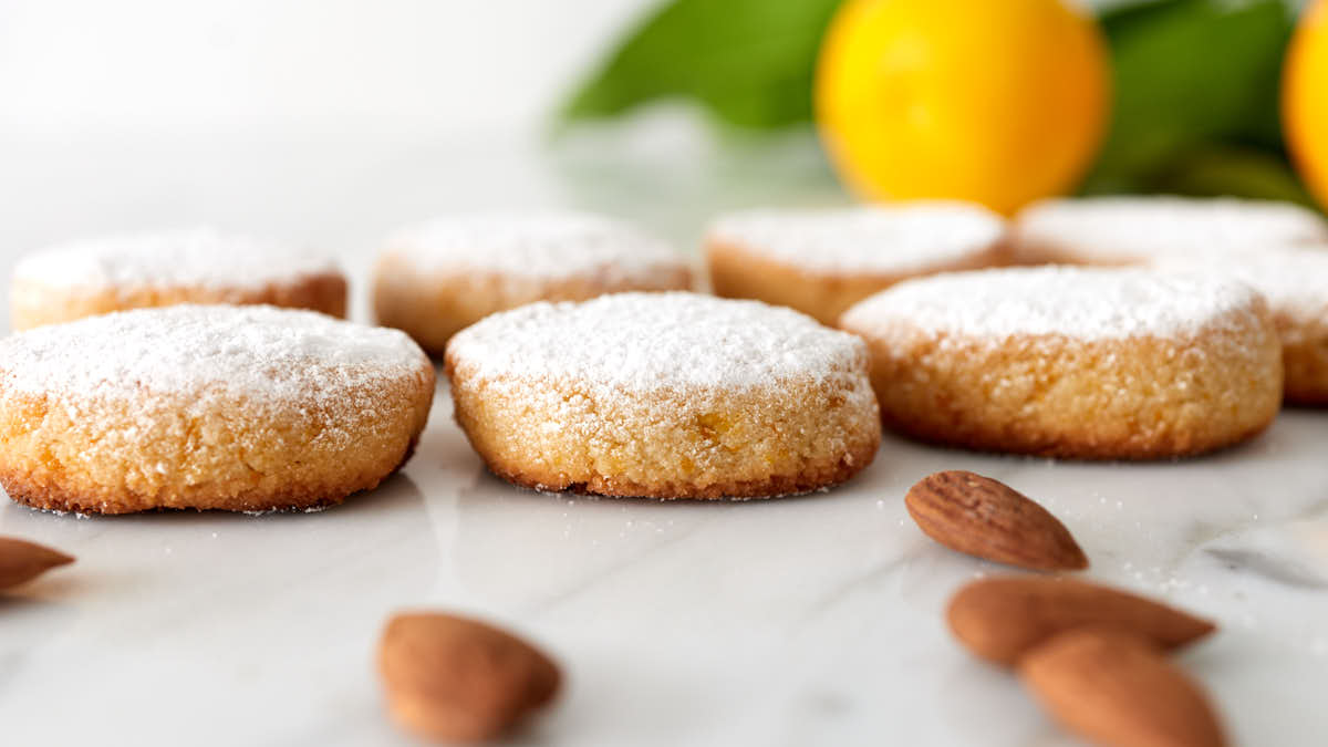 Round cookies dusted with powdered sugar are arranged on a marble surface, with whole almonds and yellow citrus fruit in the background.