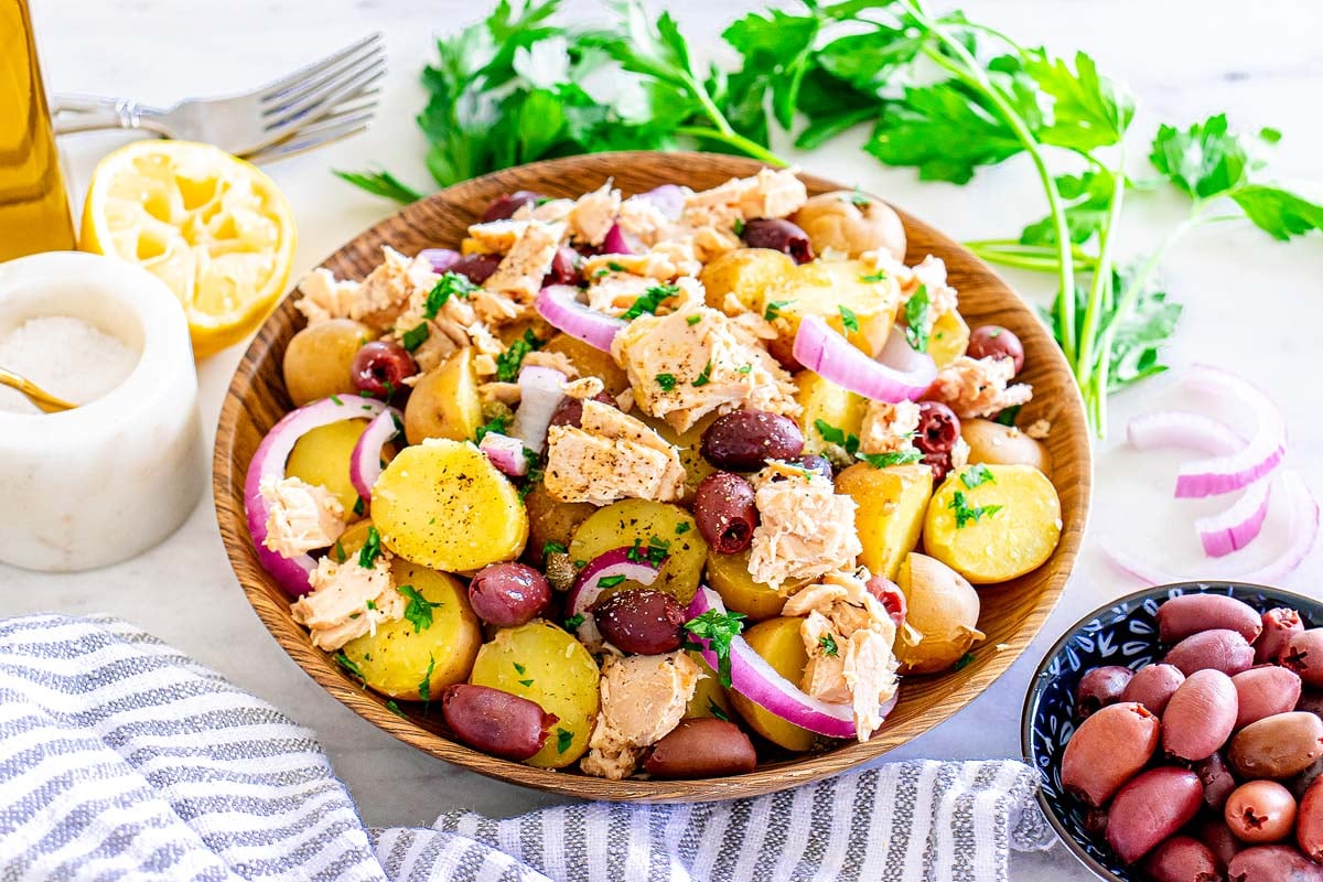 A bowl of potato salad with tuna, red onion, parsley, and kalamata olives, surrounded by fresh parsley, a halved lemon, and a bowl of olives on a white surface.