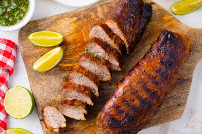 Two grilled pork tenderloins, one sliced, are displayed on a wooden cutting board with lime wedges.