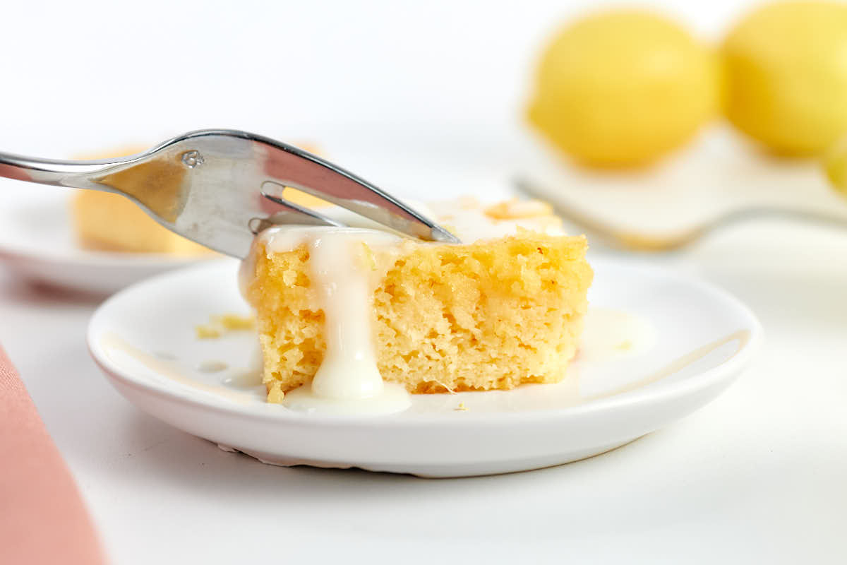 A fork cutting into a square piece of cake with white icing on a small white plate; lemons are blurred in the background.