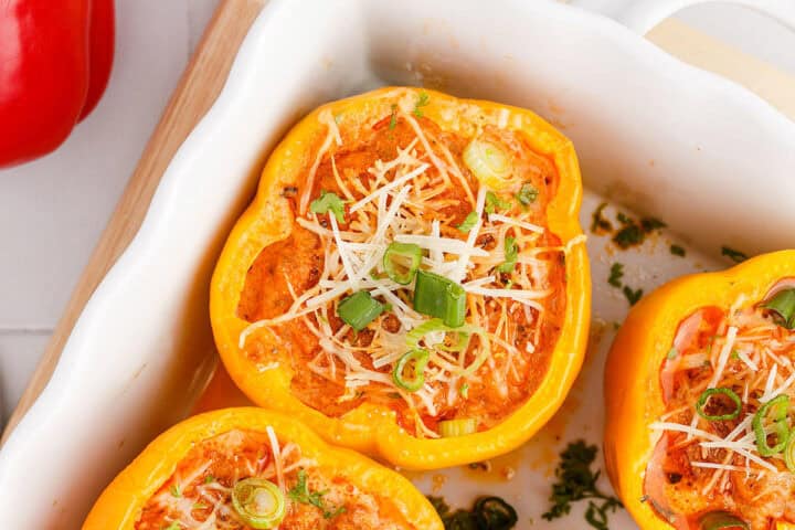 Close-up of yellow bell peppers stuffed with a filling, topped with shredded cheese and chopped green onions in a white baking dish.