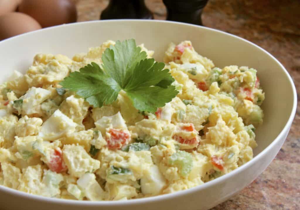 A bowl of potato salad with chopped vegetables and a parsley garnish, placed on a countertop.