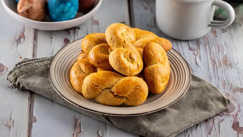 A plate of twisted, golden-brown cookies sits on a cloth napkin beside a mug and a bowl of colored eggs on a rustic wooden table.