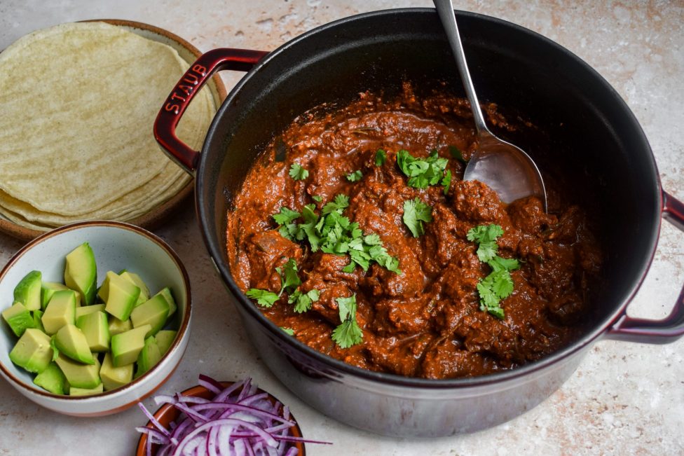 A pot of shredded beef in red sauce garnished with cilantro, with a spoon inside. Nearby are corn tortillas, diced avocado, and sliced red onions.