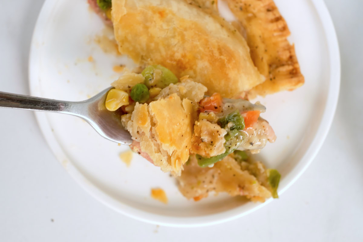 A fork holding a bite of vegetable pot pie over a white plate with flaky crust and mixed vegetables visible.