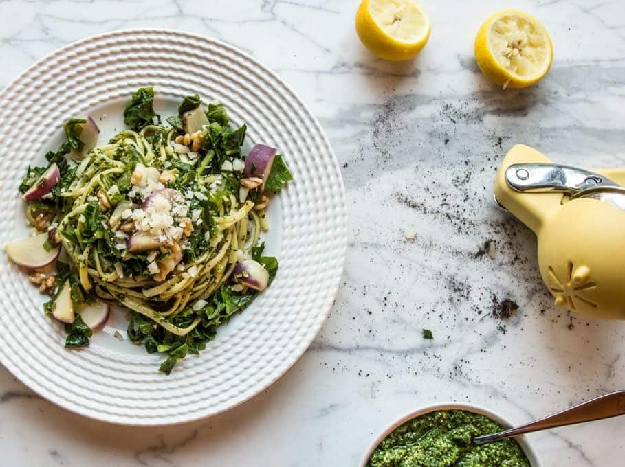 A plate of spaghetti with greens, cheese, and nuts on a marble countertop; nearby are lemon halves, a yellow citrus press, and a small bowl of green pesto.
