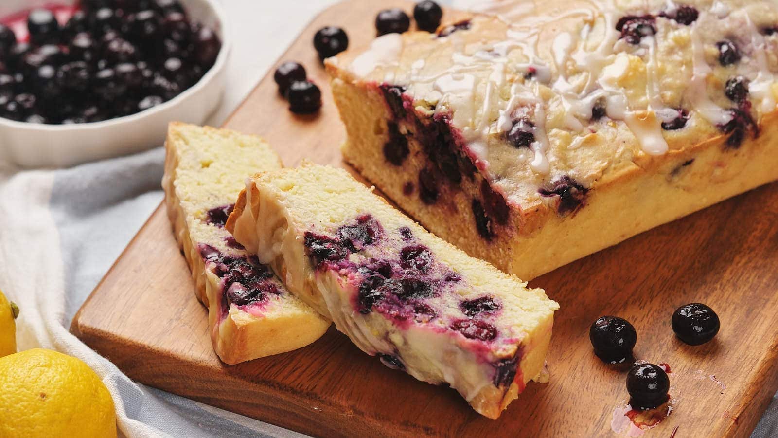 A loaf of blueberry bread with icing sits on a wooden board, with two slices cut and fresh blueberries scattered nearby.
