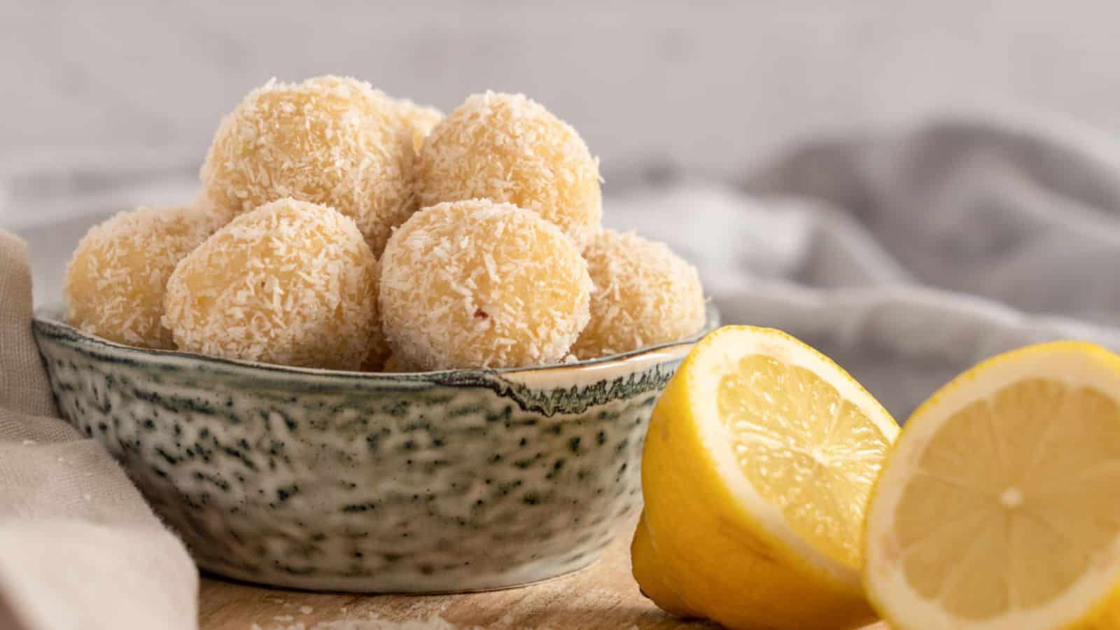 A bowl of coconut-covered lemon balls sits on a wooden surface next to two lemon halves.
