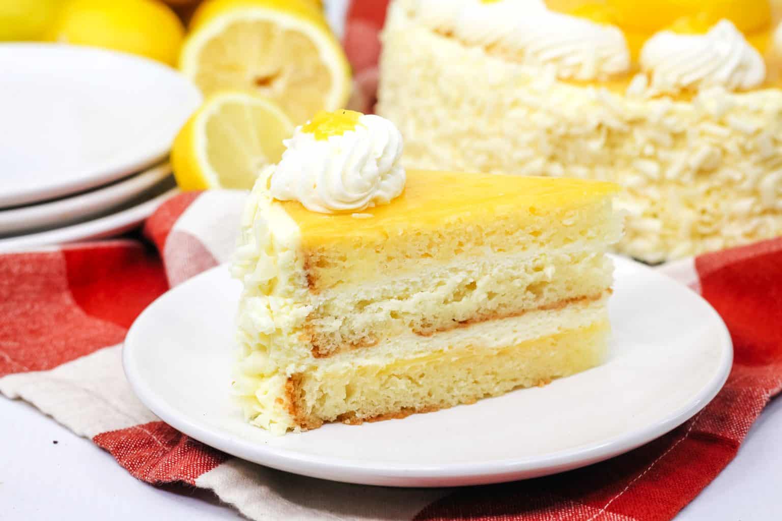 A slice of lemon layer cake with white frosting and lemon topping on a white plate, with the rest of the cake and lemons in the background.