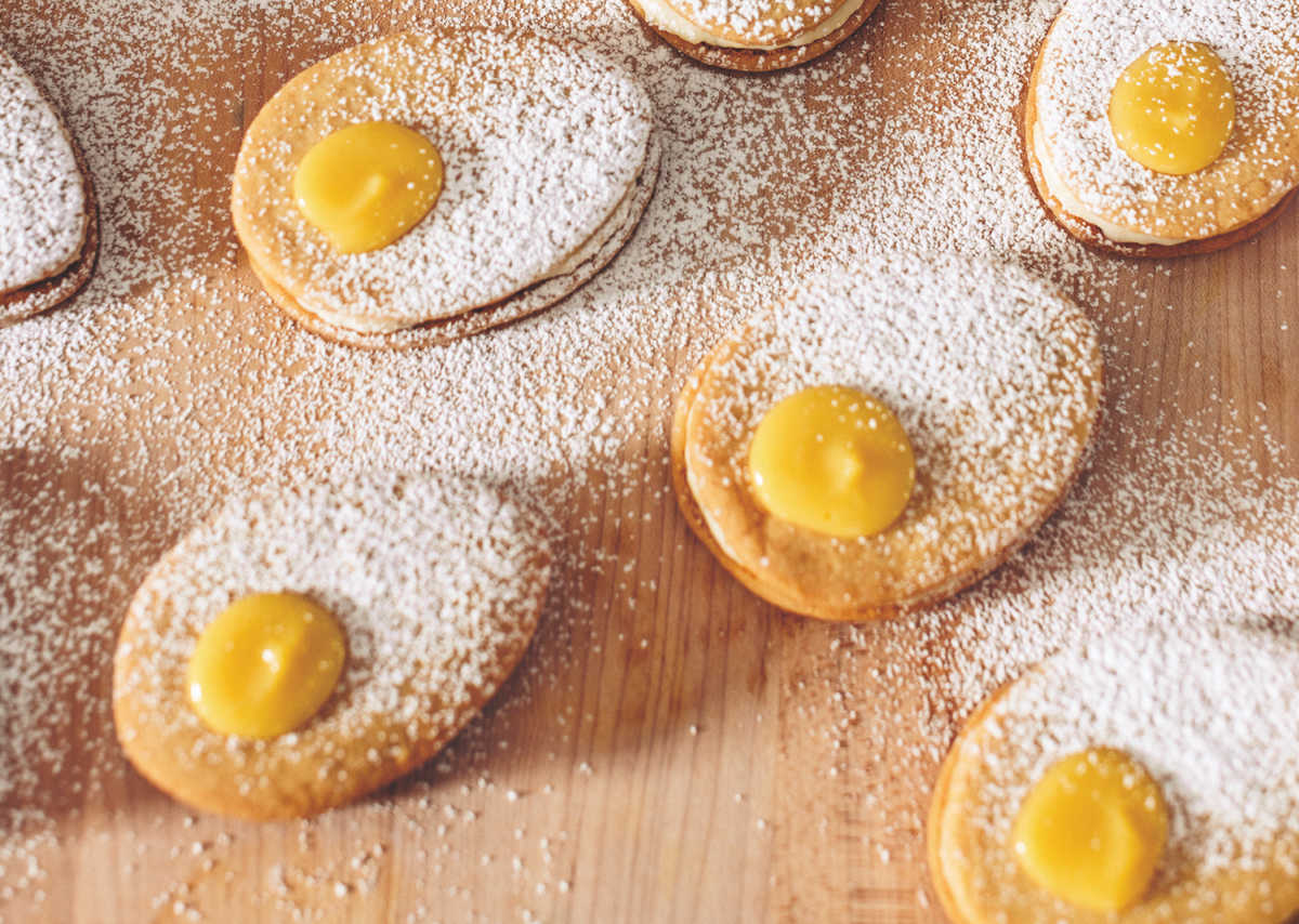 Oval-shaped cookies on a wooden surface dusted with powdered sugar, each topped with a dollop of yellow filling in the center.
