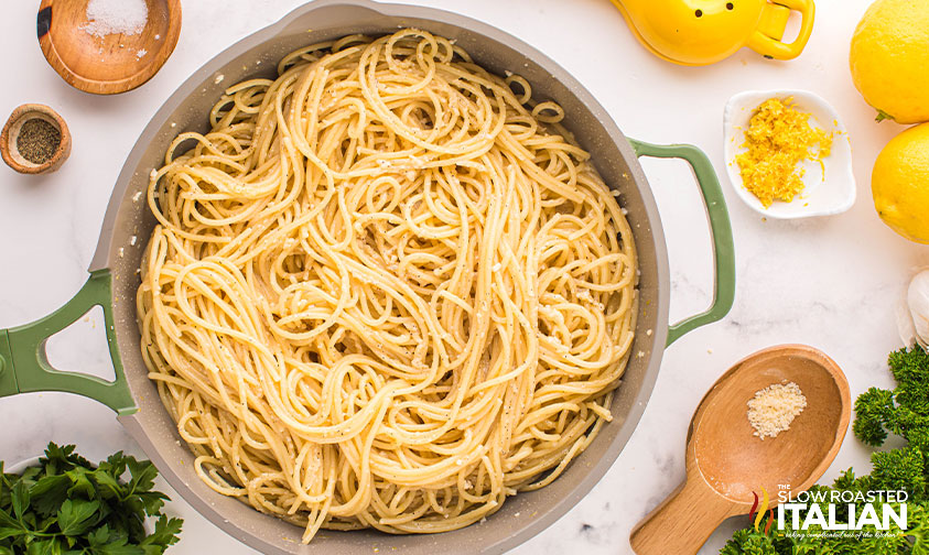 Cooked spaghetti tossed with a creamy, peppery sauce in a large green pot, surrounded by lemons, seasonings, and parsley on a white countertop.