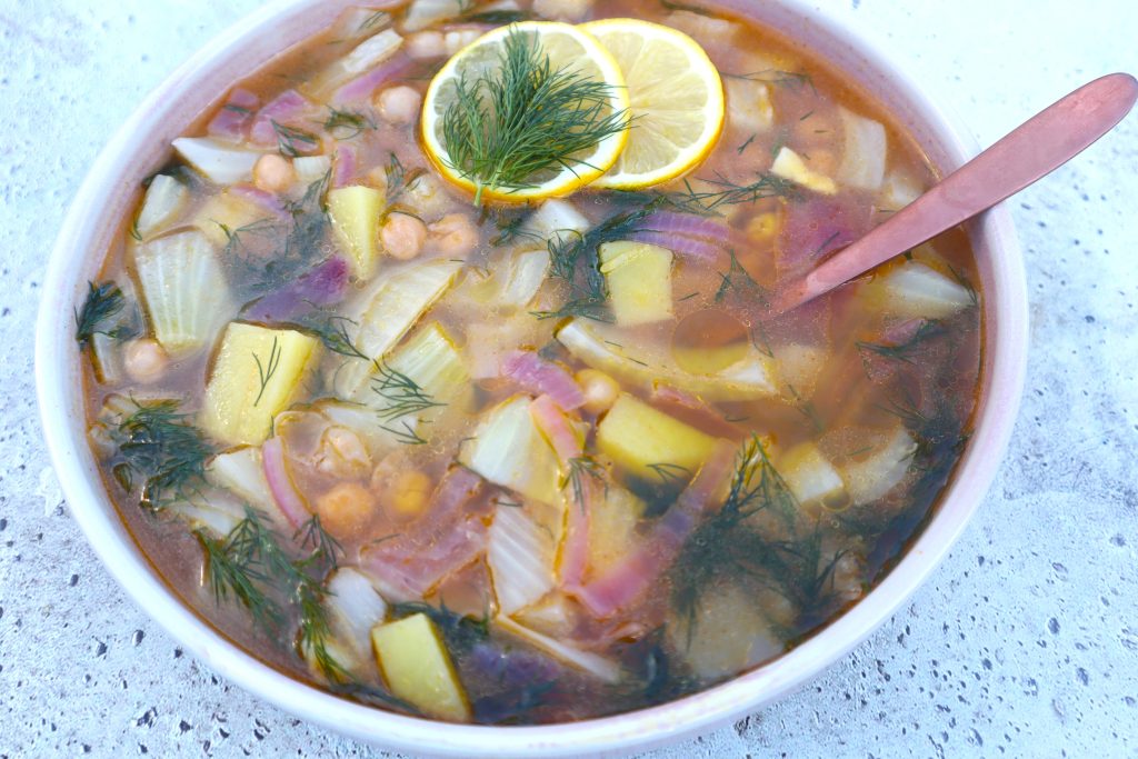A bowl of vegetable soup with chickpeas, potato chunks, red onions, fresh dill, and lemon slices on top; a spoon rests in the bowl.