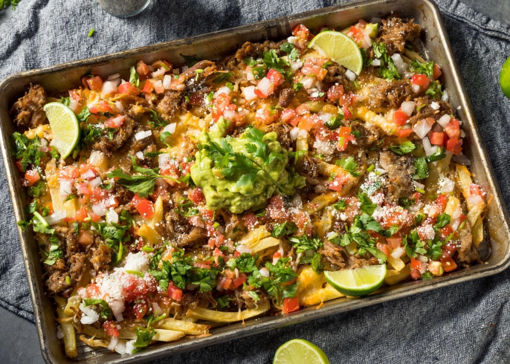 A sheet pan topped with loaded carne asada fries, melted cheese, guacamole, pico de gallo, cilantro, lime wedges, and cotija cheese.