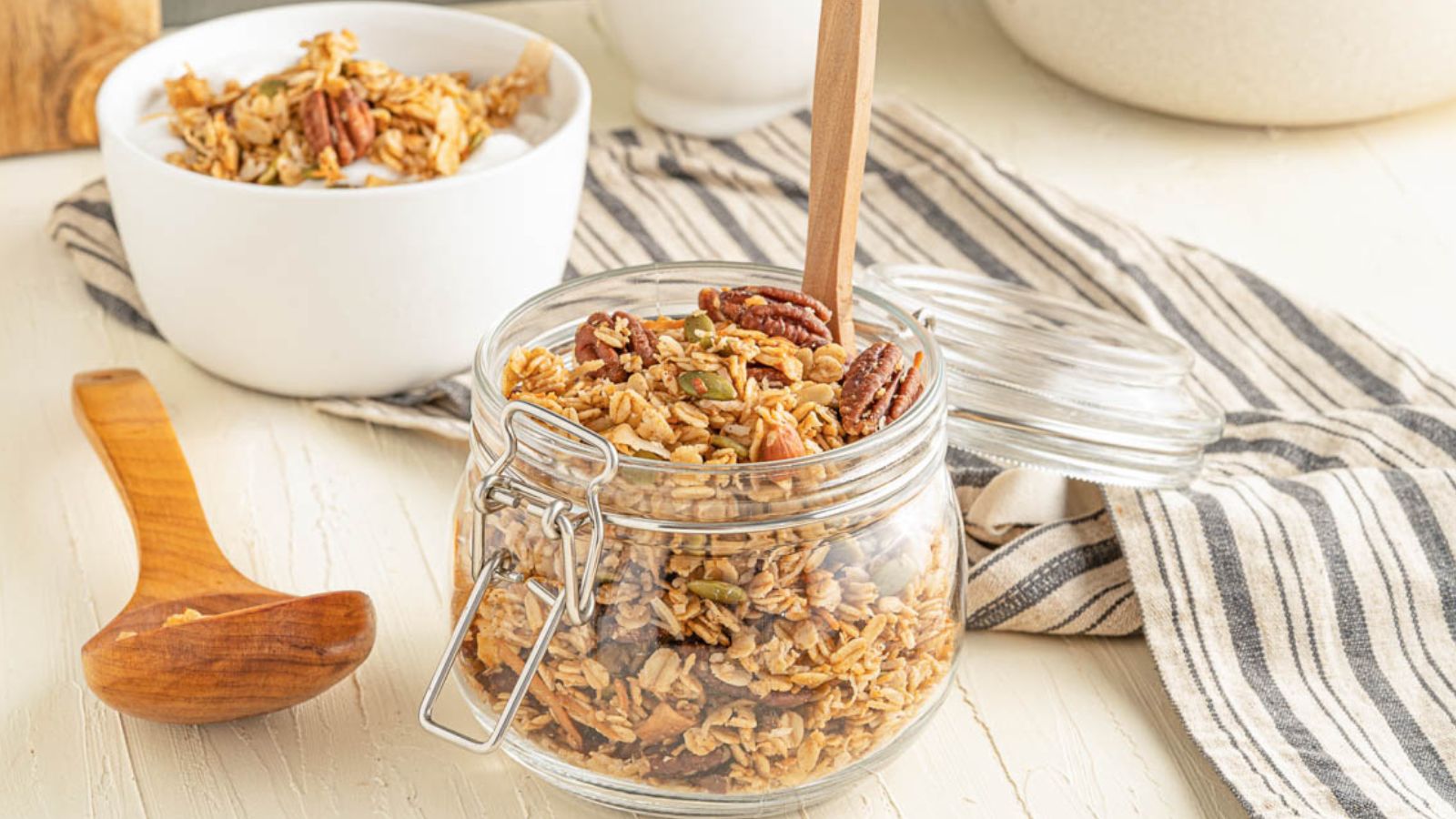 A glass jar and a white bowl filled with granola, topped with pecans, sit on a striped cloth with a wooden spoon nearby.