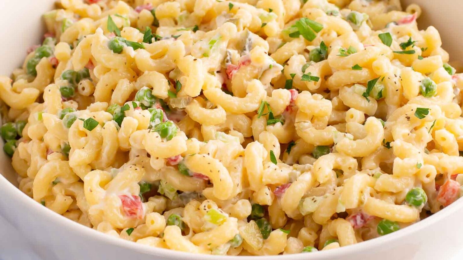 A bowl of macaroni salad with elbow pasta, peas, diced red peppers, and a creamy dressing, garnished with chopped parsley.