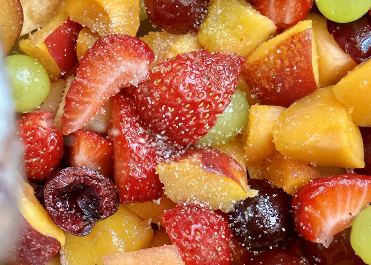 Close-up of assorted fresh fruit pieces, including strawberries, cherries, grapes, and peaches, sprinkled with granulated sugar.