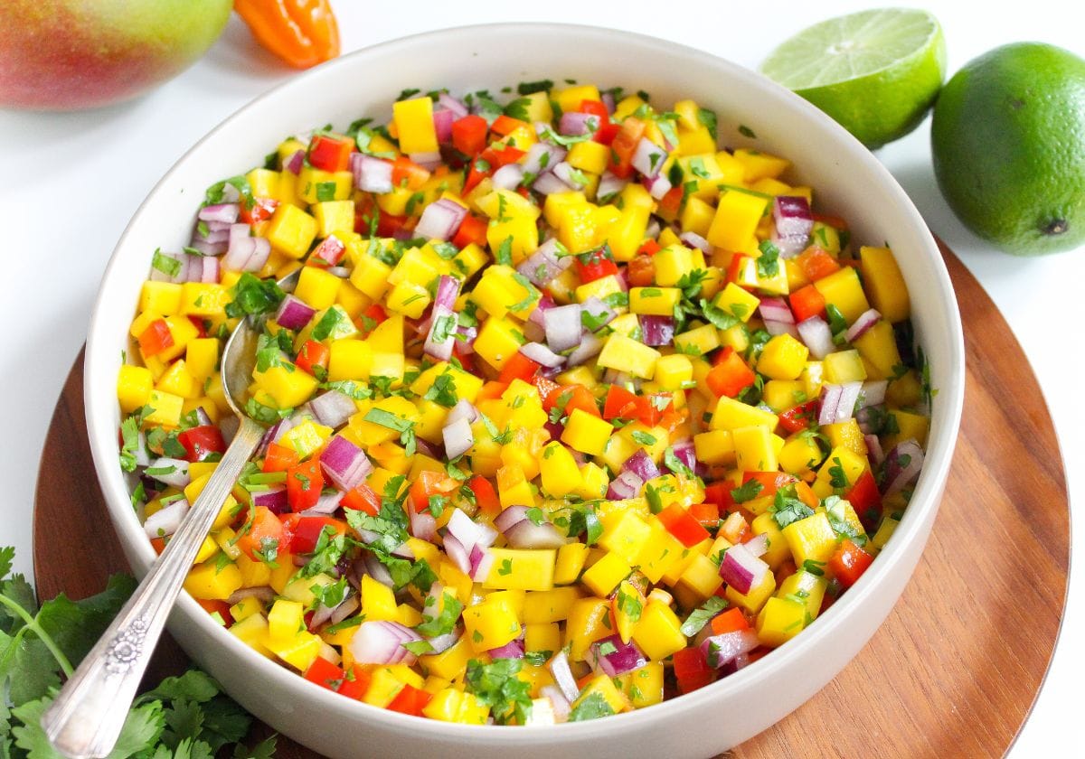 A bowl of mango salsa with diced mango, red onion, bell pepper, cilantro, and jalapeño, with a spoon in the bowl and fresh limes nearby.