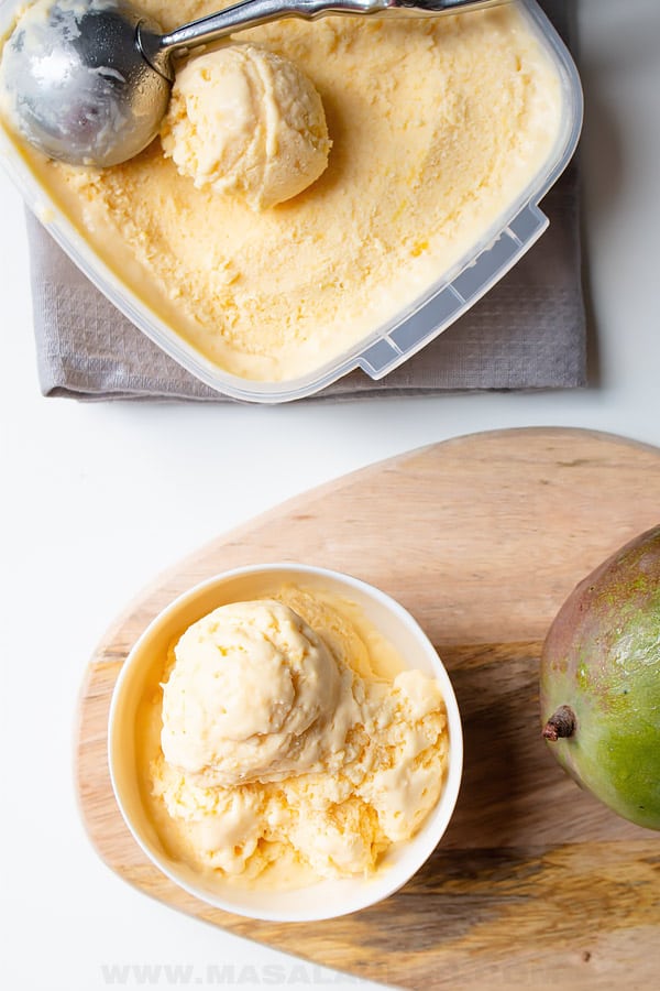 A bowl of mango ice cream with two scoops, a container of ice cream with a scoop, and a whole mango on a wooden board.