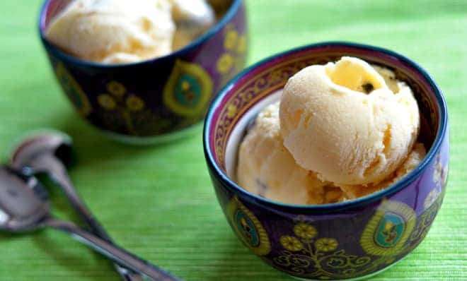 Two scoops of vanilla ice cream in a decorative bowl with another bowl and two spoons in the background on a green surface.