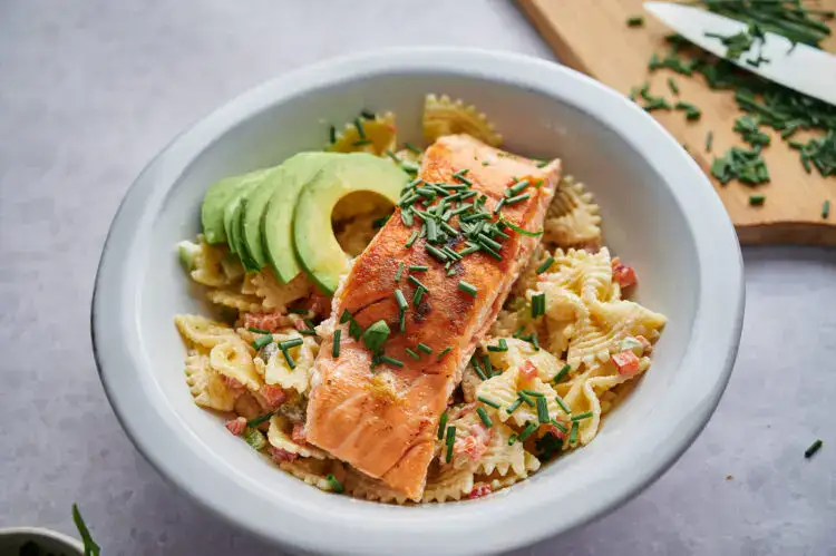 A bowl of pasta salad with rotini, cherry tomatoes, croutons, lettuce, shredded cheese, and pieces of chicken on a blue napkin.