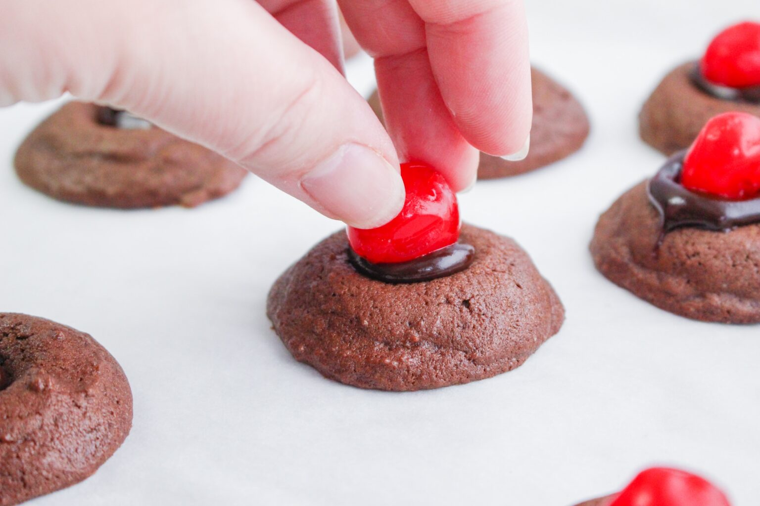 A hand places a red cherry on top of a chocolate cookie with chocolate filling, surrounded by similar cookies on a white surface.