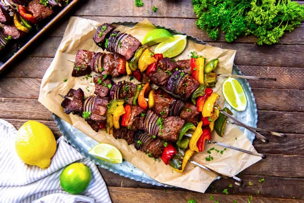 Four grilled beef and vegetable kebabs on skewers served on a tray with parchment paper, garnished with parsley and accompanied by lime and lemon wedges.