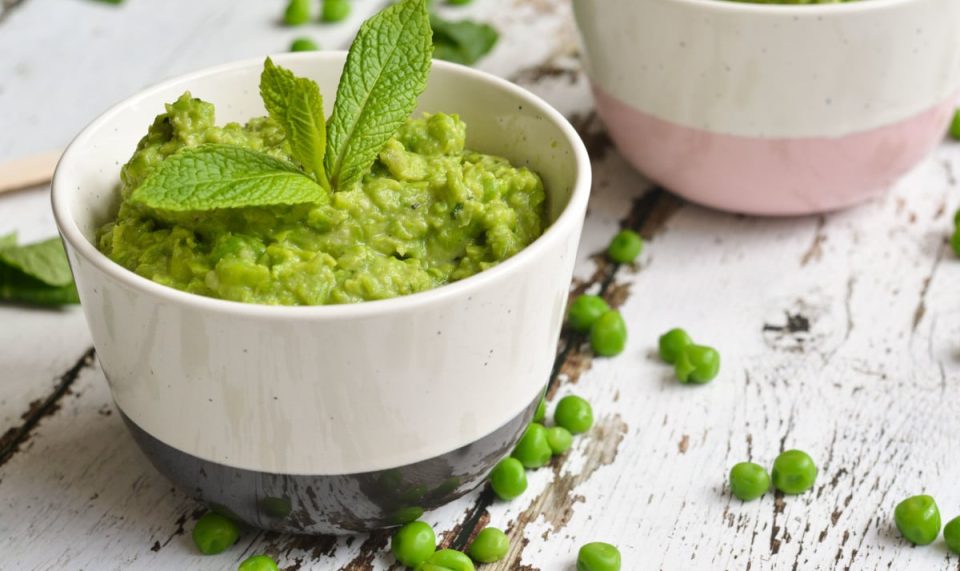 A ceramic bowl filled with green mashed peas and garnished with fresh mint leaves sits on a rustic white wooden surface.