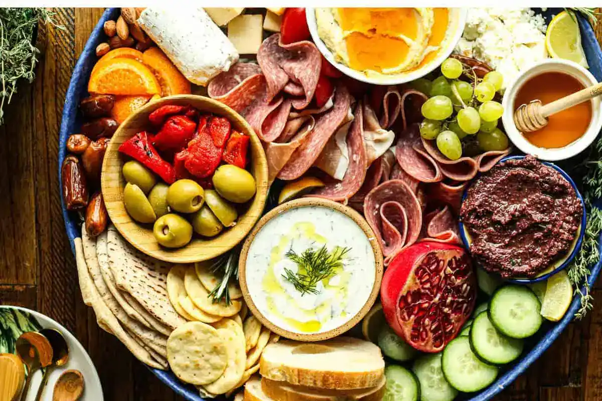 A charcuterie board with assorted meats, cheeses, crackers, pita, olives, dips, roasted peppers, grapes, cucumber slices, pomegranate, and honey.