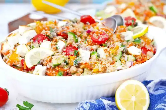 A white dish filled with a colorful salad containing quinoa, cherry tomatoes, cucumber, feta cheese, lemon slices, and herbs, with serving utensils and a cut lemon beside it.