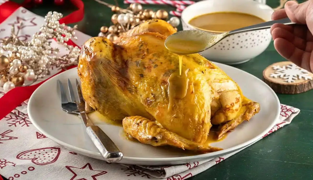 A whole roasted chicken is served on a white plate with gravy being poured over it, accompanied by a fork and knife, festive decorations, and a bowl of sauce in the background.